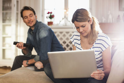 Woman using laptop while man looking at her in house