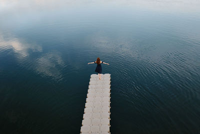 High angle back view of female standing with outstretched arms on quay near lake and enjoying freedom