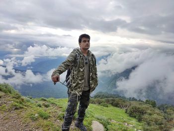 Young man standing by cliff against cloudscape