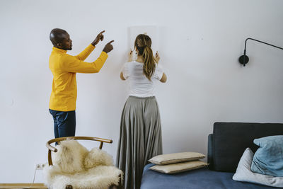 Mid adult man guiding girlfriend adjusting painting on wall at home