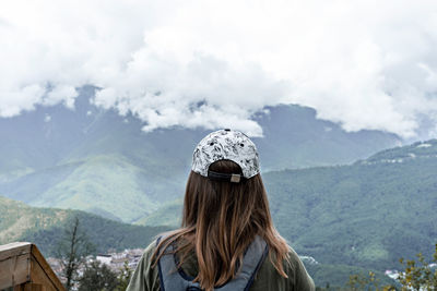 Rear view of woman in cap with backpack looking at view of mountains and cloudy sky on  viewpoint