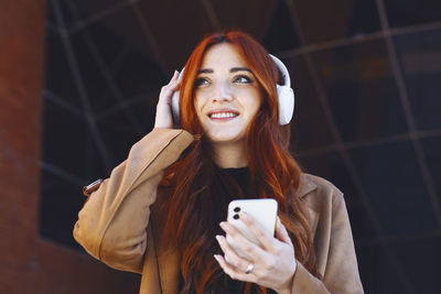 Young redhead woman smiling, while listening to music with wireless headphones and smartphone,