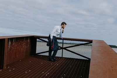 Young man looking away while standing by railing against cloudy sky