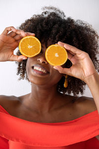 Black woman with afro hair holding cut orange halves in front of her eyes having fun