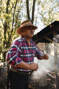 Argentinian male in hat sharpening knife between smoke diffusing in air in countryside