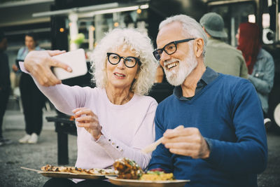 Senior woman taking selfie with smiling man eating meal against food truck in city