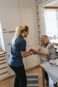 Side view of female caregiver holding hands while talking with senior woman at home