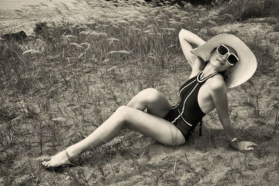A woman in a swimsuit, hat and sunglasses sunbathes in summer on the riverbank among the grass
