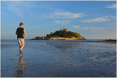 Rear view of young man standing on beach against sky in front of st michaels mount