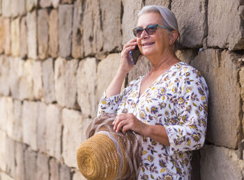 Smiling senior woman talking on phone standing by stone wall outdoors