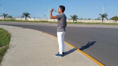 Young man drinking water while standing on footpath against clear sky during sunny day