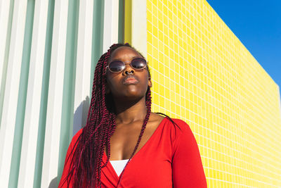 From bellow young african american female in trendy outfit and glasses looking away while leaning against colorful wall on a sunny day
