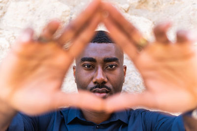 Serious african american adult guy looking at camera while demonstrating triangle shape with hands near fence in street in daylight