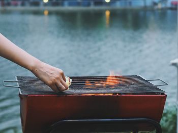 Cropped hand of woman holding food over barbecue grill by lake