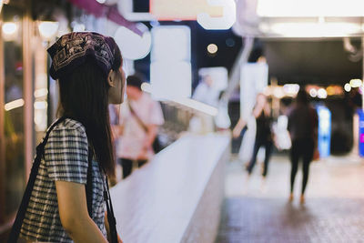 Side view of woman sitting on wall by footpath in illuminated city