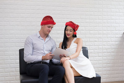 Couple using digital tablet while sitting on seat against white brick wall