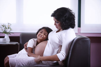 Smiling daughter with pregnant mother in living room at home