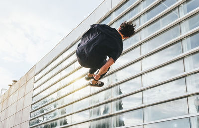 From below back view of anonymous male in casual clothes jumping and performing parkour trick in air on urban street with modern buildings