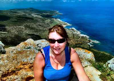 High angle portrait of woman wearing sunglasses sitting on mountain against sea