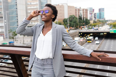 Young smiling black female manager wearing trendy outfit and eyeglasses leaning on railing while resting in balcony of modern building