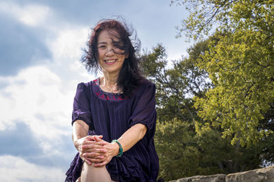 Portrait of smiling senior woman sitting outdoors against tree