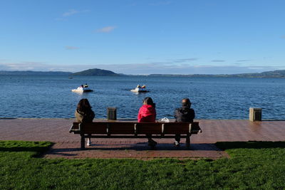 Rear view of people sitting on bench at promenade by lake against sky