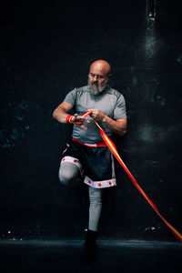Muscular mature male bearded athlete in sportswear putting on strap on wrist while standing against dark wall and preparing for training in gym