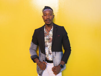 Serious african american male model in fancy outfit standing near yellow wall on street and looking at camera