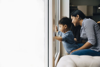 Side view of mother and son looking through window while sitting at home