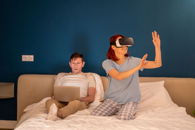 Interested woman in glasses virtual reality sits on bed with husband manages vr goggles from laptop