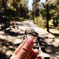 Cropped hand holding gold ring over railroad tracks during sunny day