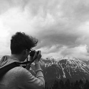 Man photographing snowcapped mountains through camera against cloudy sky