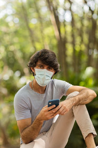 Young man wearing a mask and a smartphone at day time at a green park . mobile phone, technology