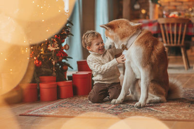 Candid authentic happy little boy in knitted beige sweater hugs dog with bow tie at home on xmas