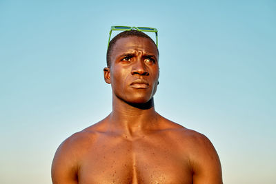 From below serious thoughtful shirtless african american male with green sunglasses on head looking away against sea and cloudless sundown sky