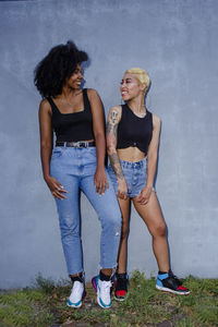 Two smiling friends lean together against a wall in matching clothes