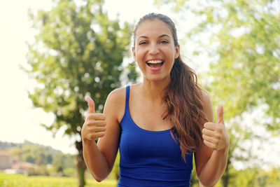 Portrait of smiling woman gesturing thumbs up while standing against trees