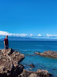 Full length of man looking at sea while standing on rock against blue sky