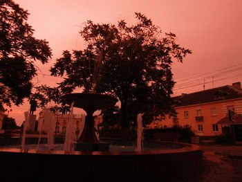 Silhouette tree by illuminated building against sky at sunset