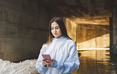 Young woman using phone while standing against wall
