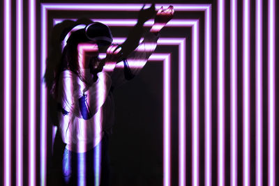 Young woman wearing virtual reality simulator standing against illuminated background