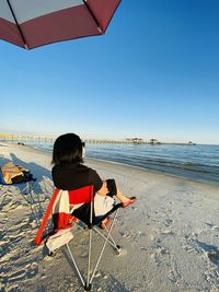 Rear view of woman sitting on chair at beach against clear sky