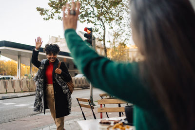 Hispanic female waving hand to excited african american friend while sitting at table with desserts in street cafe