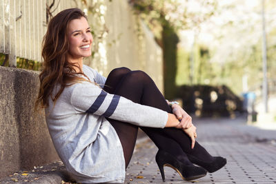 Side view of cheerful young woman sitting on footpath