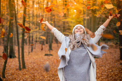 Smiling young woman with arms outstretched standing amidst falling leaves during autumn