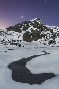 Picturesque scenery of cold river flowing among snowy valley leading to high rocky mountain under starry sky in dusk