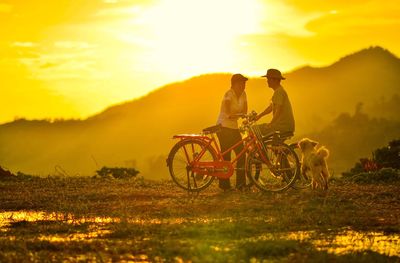 Man and woman with bicycles and pet on field against sky during sunset