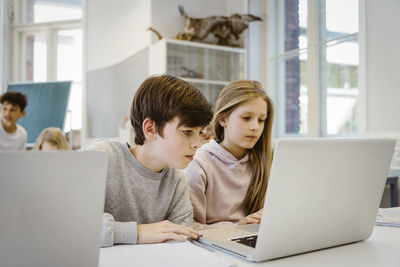 Concentrated boy watching laptop with female friend at desk in classroom