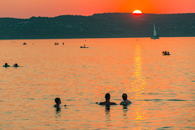 Silhouette people swimming in sea against orange sky