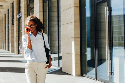 Young smiling businesswoman talking on smart phone while walking in corridor